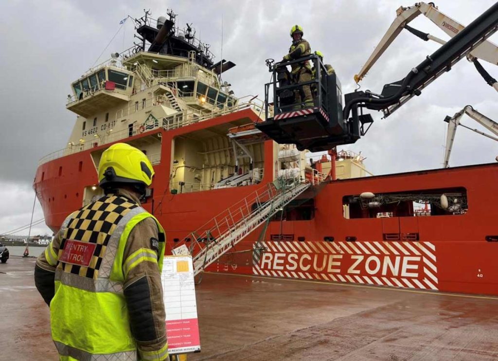 A Scottish Fire and Rescue Service Entry Control Officer looking on to two firemen on an aerial platform in front of the Atlantic Kestrel vessel during a safety exercise at Montrose Port Authority
