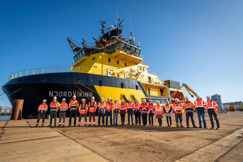 Montrose Port Authority Team standing in single file on the quayside in orange PPE and white hard hats standing in front of the Njord Viking vessel