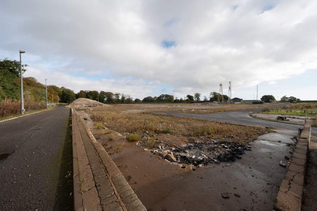 The empty industrial site at Montrose Commercial Park site which shows the site mostly empty with overgrown weeds and some debris. 