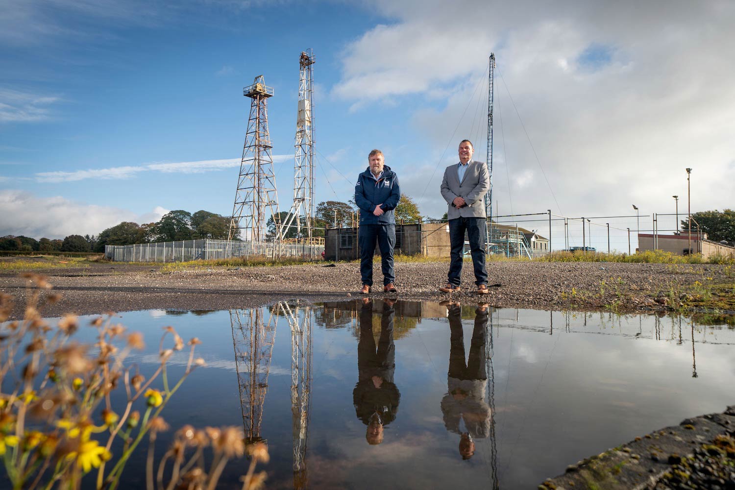 Montrose Port CEO Tom Hutchison and Equipco Investments Ltd owner Mark Stephens standing together at Montrose Commercial Park. They're reflection is show in a puddle infront of them with some offshore energy equipment for training behind them.