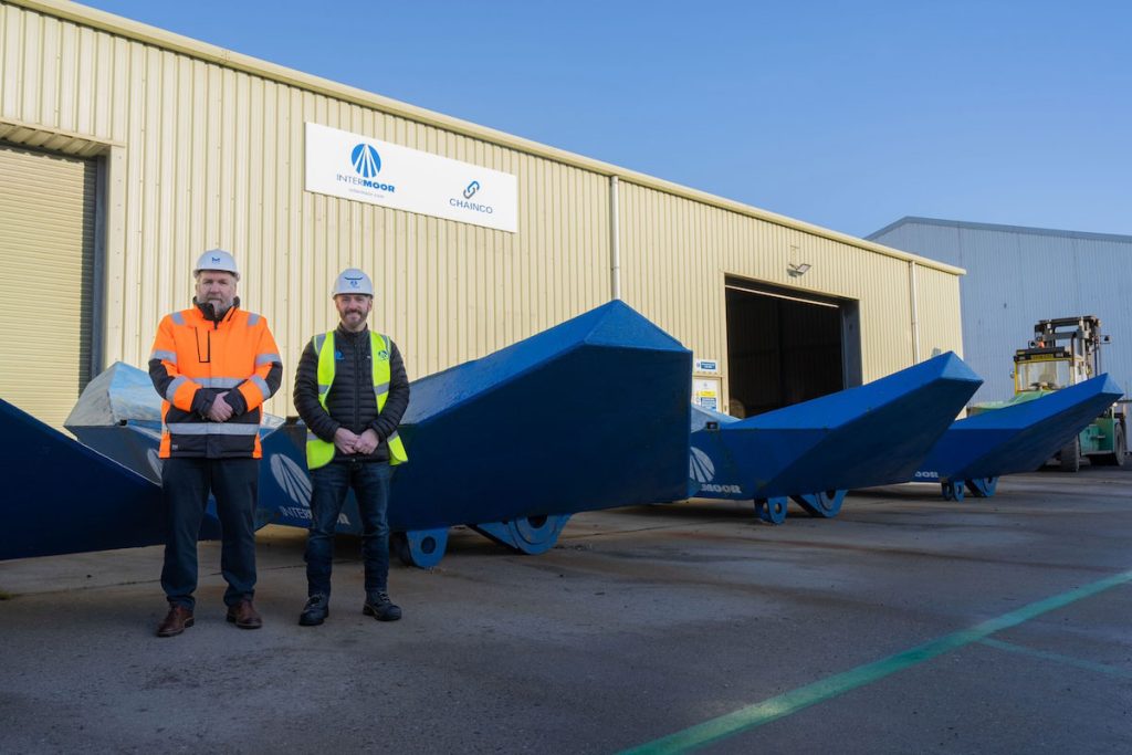 Tom Hutchison, CEO of Montrose Port, with Frank Burns, General Manager at InterMoor UK, both standing in PPE and hard hats in front of InterMoor branded anchors and warehouse.