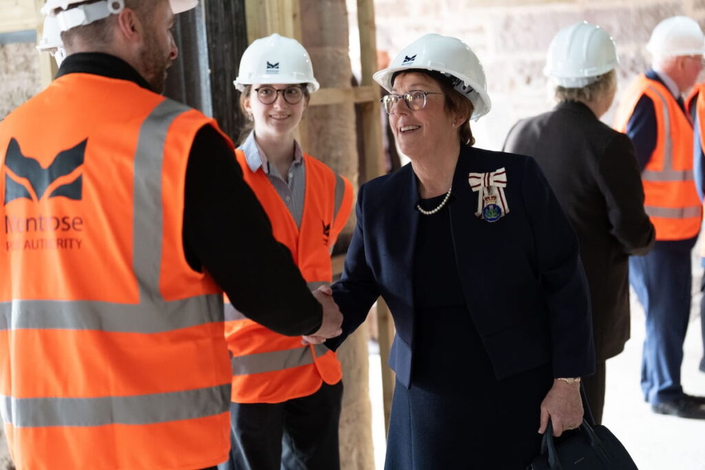 A woman in a dark suit and white hard hat shakes hands with a man in an orange safety vest at Montrose Port during the Duke of Edinburgh’s visit; other people in safety gear are visible in the background.