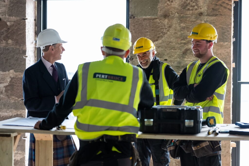 Four men wearing hard hats and high-visibility vests stand around a worktable at a construction site. One man, in a suit and white hard hat, speaks to three workers in yellow helmets. Construction tools and papers are on the table.