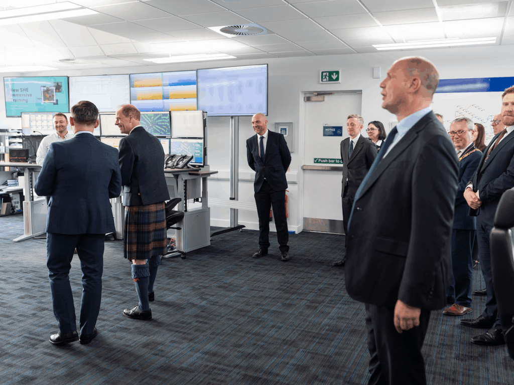 A group of men in suits and one in a kilt stand in a control room with computer monitors on desks and screens on the walls. Most people are smiling or watching something off camera.