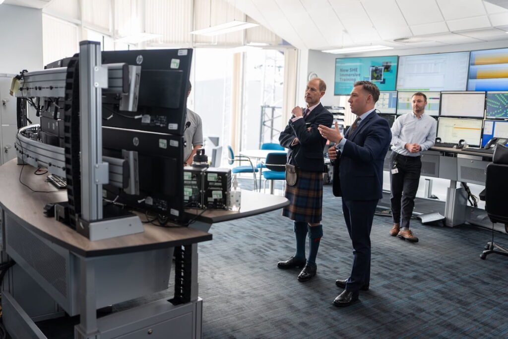 Several men stand and talk in front of large computer monitors in a modern control room. One man is wearing a kilt, while others are in business attire. Screens and office equipment are visible in the background.