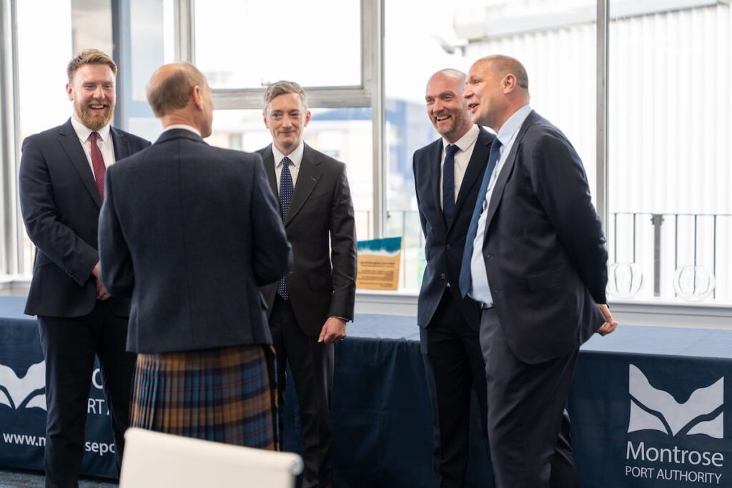 Five men in suits stand talking and smiling in a room with large windows. Two tables with Montrose Port Authority banners are in the background. The Duke of Edinburgh has his back to the camera and is wearing a kilt.