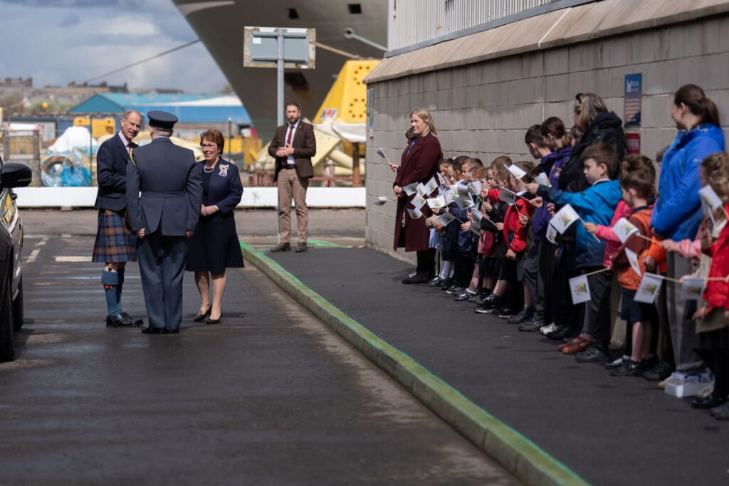 A group of adults in formal attire greet each other on a paved area near a building whilst children and adults stand in a queue nearby, holding small flags and watching the scene.