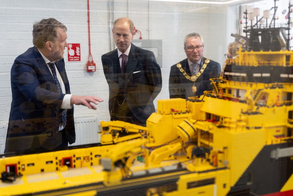 Three men in formal attire stand behind a glass display case, observing a large yellow and black model ship. One man gestures towards the model whilst the others listen. A fire alarm and red pipes are visible on the wall behind them.