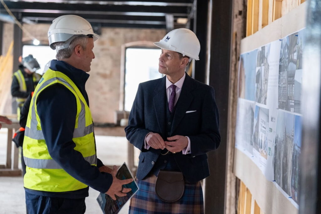 Two men wearing hard hats stand indoors at a construction site, having a conversation. One wears a high-visibility vest, and the other wears a suit jacket and kilt. Construction plans are displayed on the wall beside them.