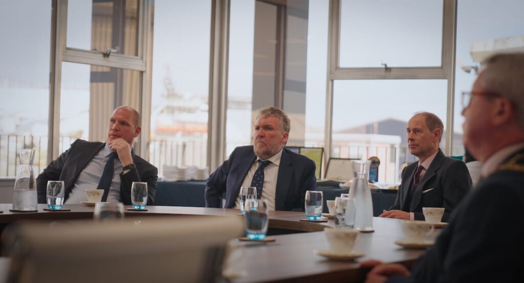 Four men in suits sit at a round table set with cups, saucers, and glasses of water in a meeting room with large windows and an outdoor view in the background.