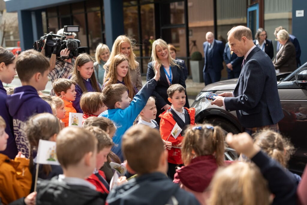 A group of school children gathers outside near a building, some holding small flags, while the Duke of Edinburgh dressed in a suit and kilt holds a gift that the school children have given him. Several adults and a camera crew are present in the background.