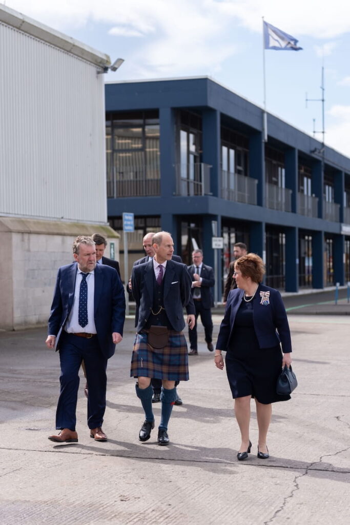 A group of formally dressed people, including a man in a kilt, walk and talk outside an industrial building. A flag is flying from the roof, and others follow behind them.