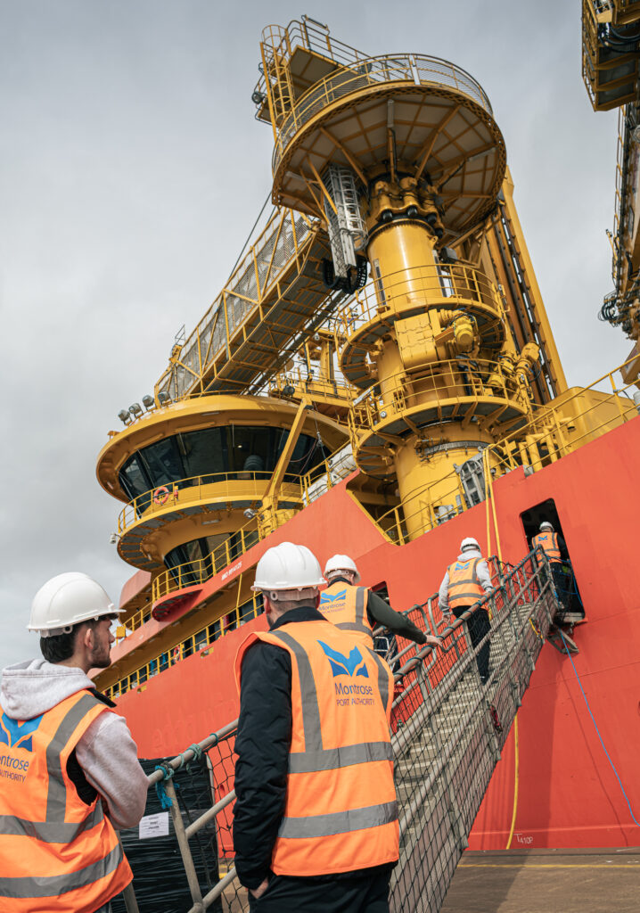 Several people wearing hard hats and Montrose Port Authority vests walk up a gangplank onto a large red and yellow industrial ship, with complex machinery visible under a cloudy sky.