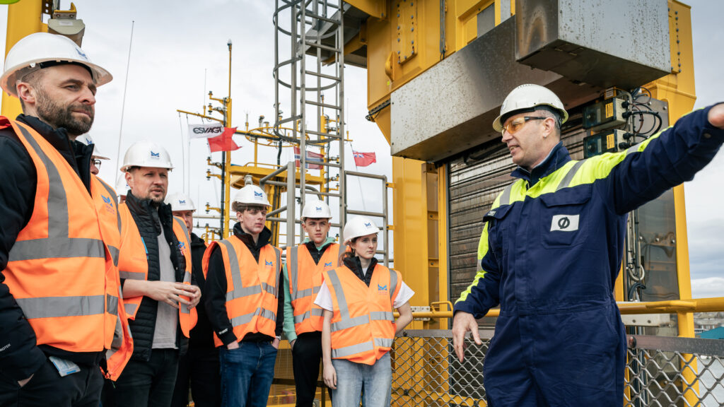 A man in a blue and yellow safety uniform and hard hat gestures while speaking to a group of people in orange safety vests and hard hats on an industrial site with metal structures.