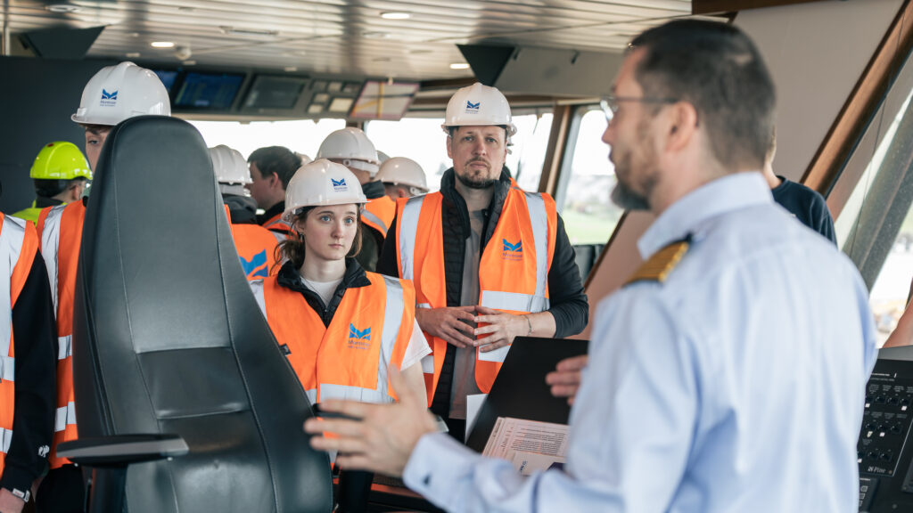 A group of people wearing hard hats and orange safety vests listen to a person in a uniform speaking inside a control room with large windows.