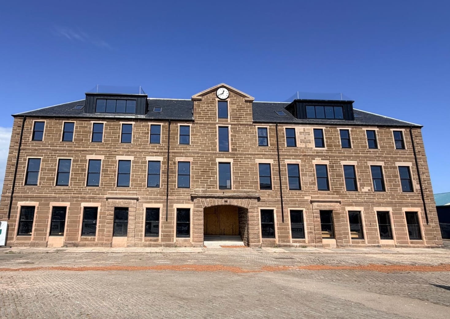A large, three-storey stone building with many windows, a central archway entrance, and a clock above the doorway. The building is set against a clear blue sky on a paved open area.