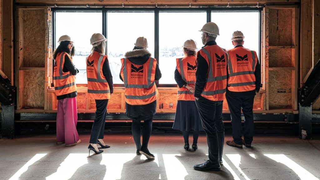 Five people wearing orange PPE vests and white hard hats stand inside a partially built structure, facing a large window opening with a view outside.