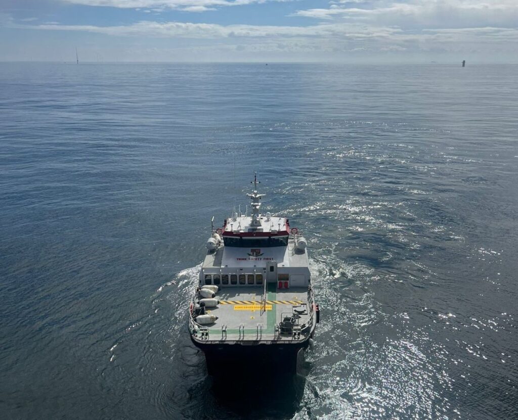 A white crew transfer vessel on calm, open water under a partly cloudy sky. The vessel creates a gentle wake as it moves away from the camera, with land barely visible on the horizon.