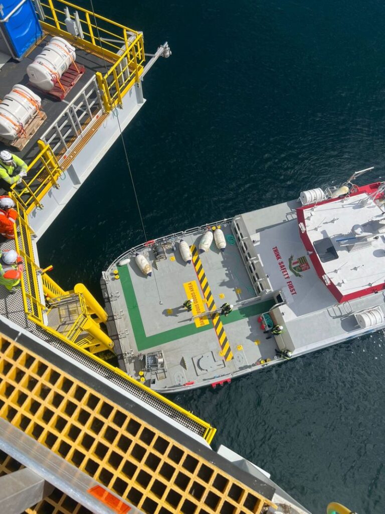 A red and white service boat is moored beside an offshore platform. Several workers in safety gear are visible on the platform, looking down at the vessel. The water is dark and calm.