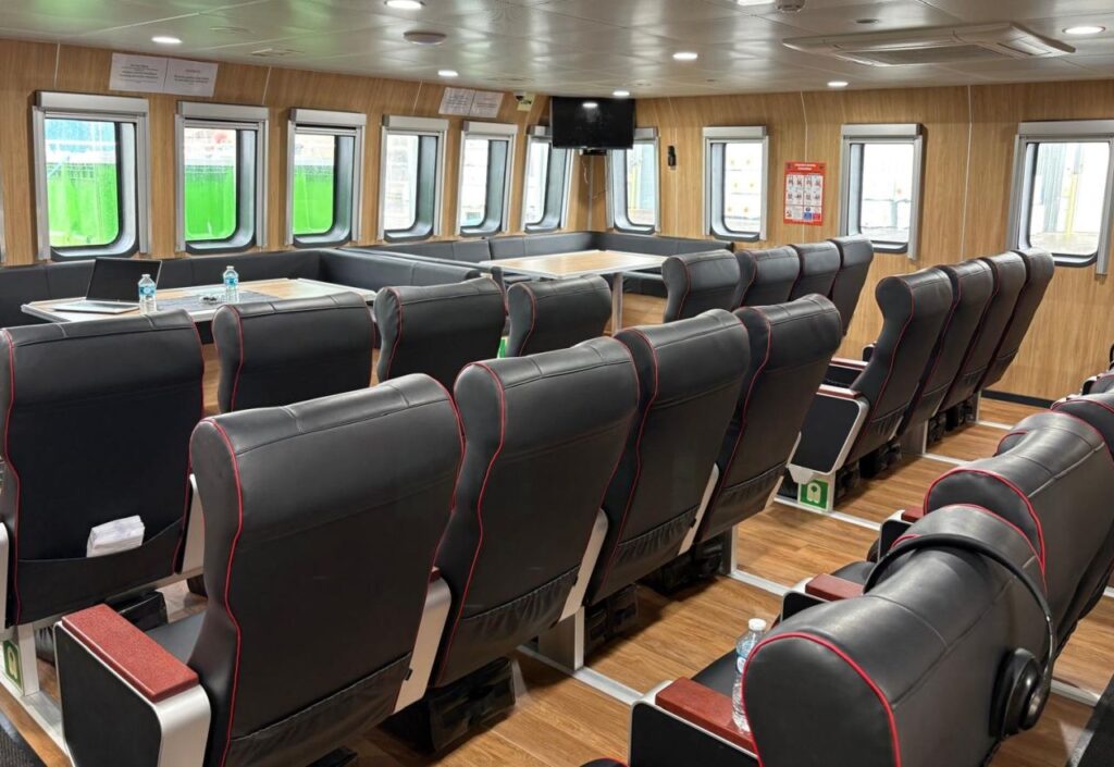 Rows of black cushioned seats with red trim are arranged in a passenger cabin with wooden panelled walls, large windows, folding trays, a TV screen, and bottled water placed on some trays.