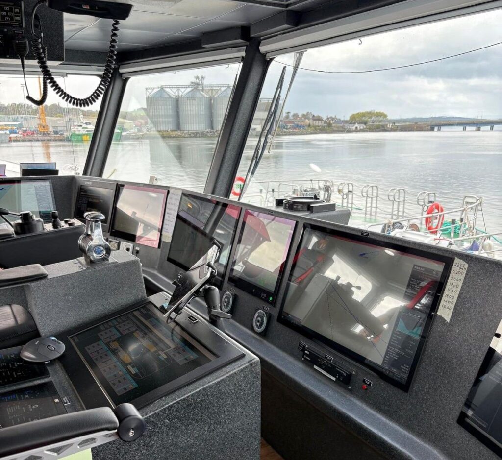 A modern ship's bridge with multiple digital navigation screens, control panels, and communication equipment, overlooking a harbour with boats, water, and industrial buildings visible through large windows.