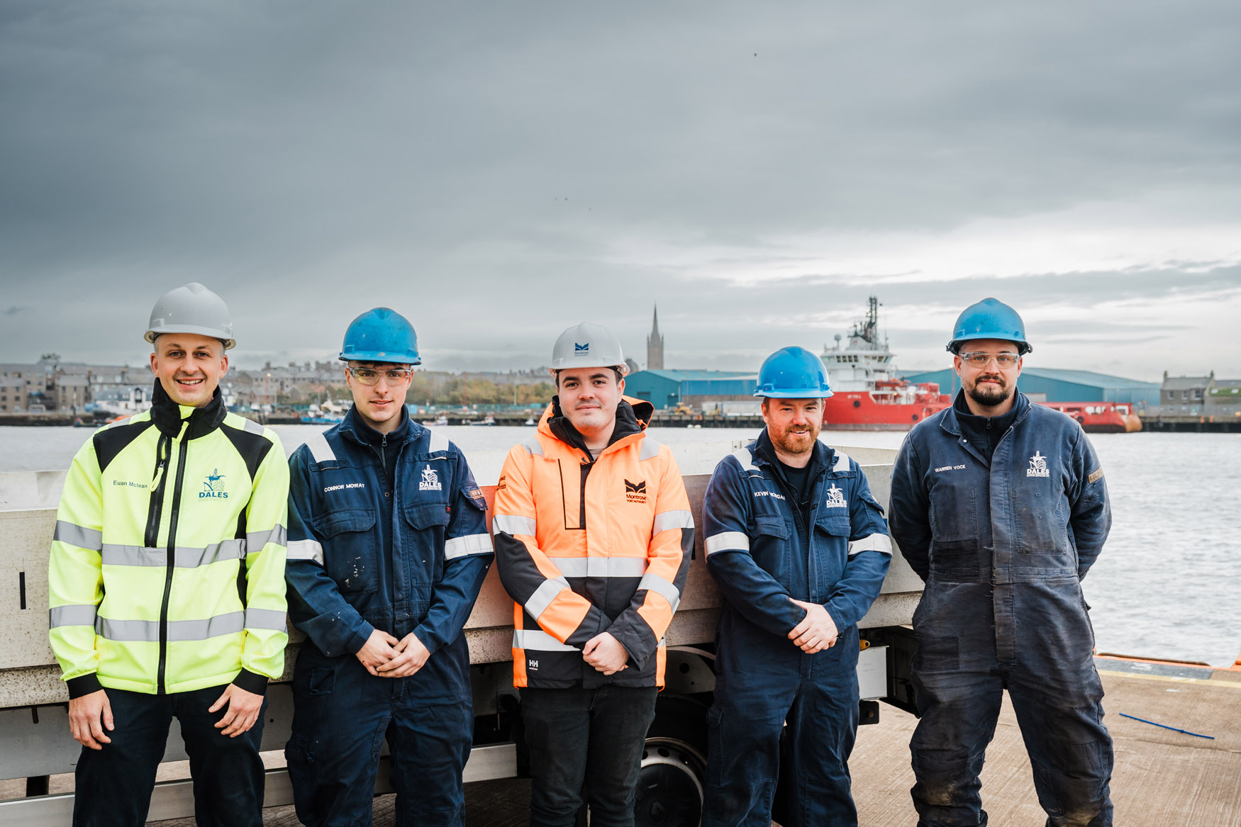 Five workers wearing safety helmets and workwear stand in a row on a quay, with industrial buildings, ships, and water visible in the background under a cloudy sky.