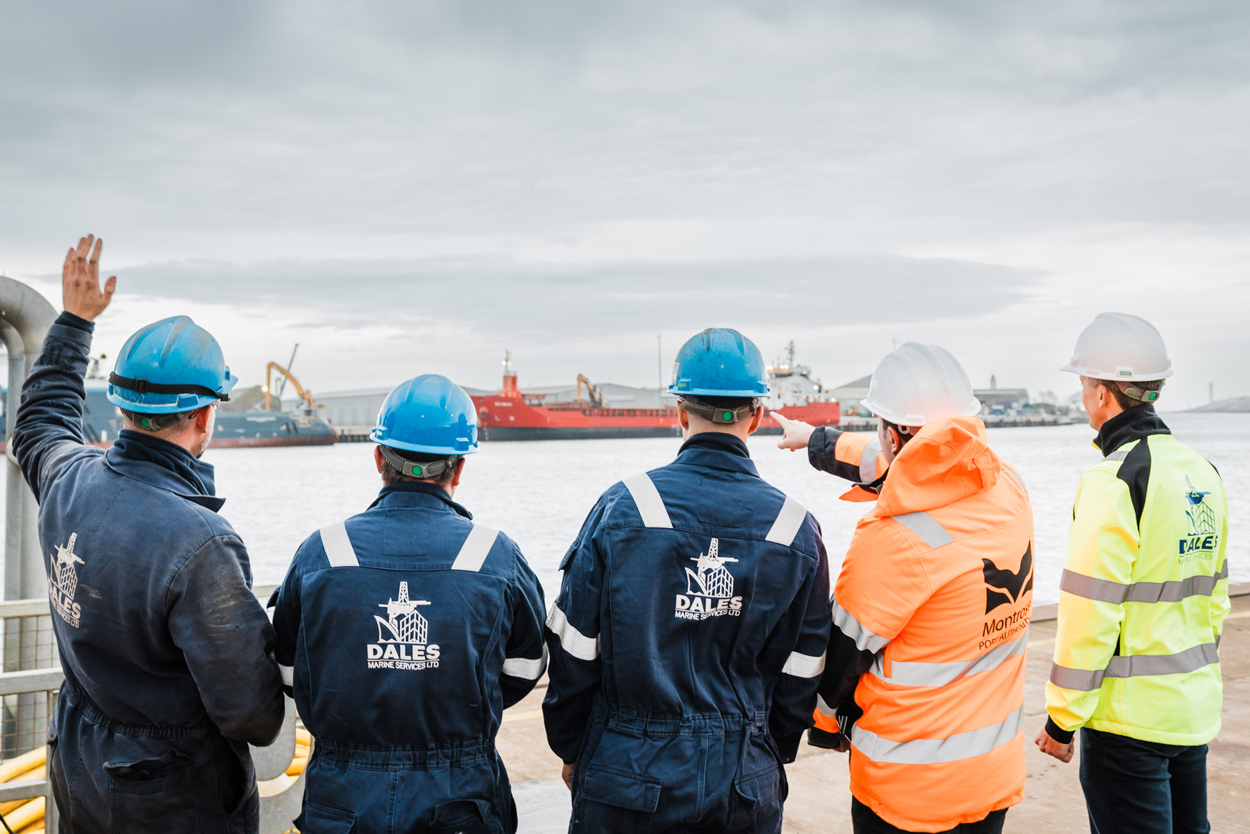 Five workers in safety gear and helmets stand on a quay, facing the water. One person points towards cargo ships in the distance, while another raises a hand. Industrial equipment and cranes are visible on the ships.