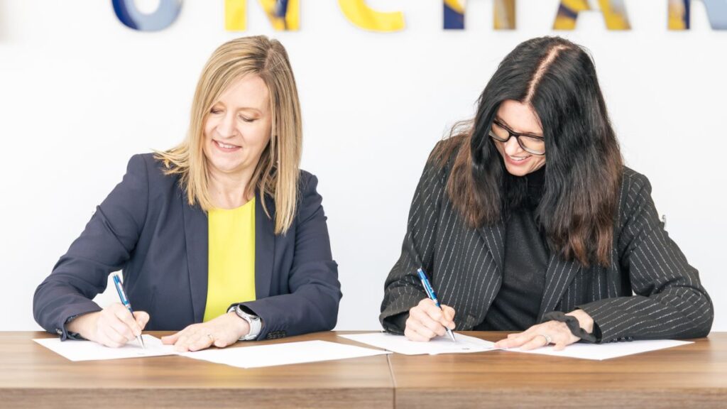 Two women sitting at a table, both smiling and signing documents with pens. They are wearing business attire and appear to be participating in a formal agreement or contract signing.