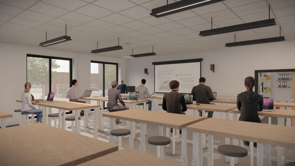 A classroom with students sitting on stools at wooden tables, facing a teacher and a whiteboard with a projector screen; some students have laptops open, and large windows are on one side of the room.