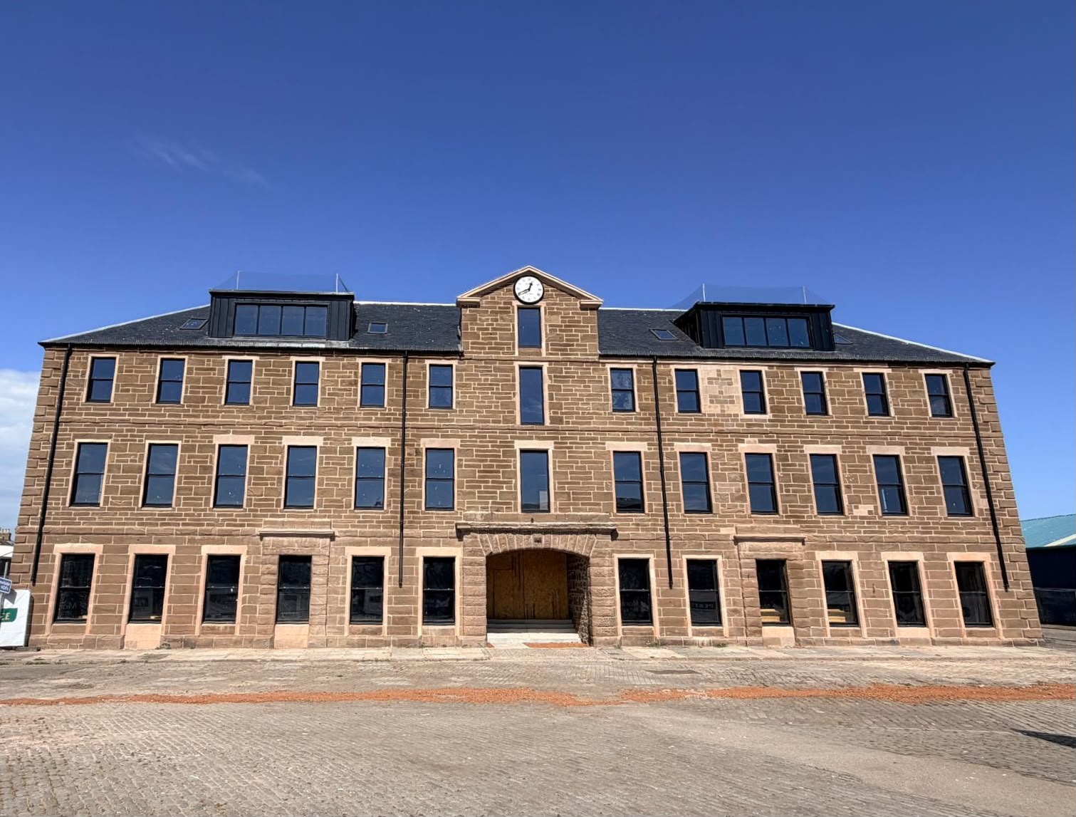 A large, rectangular, three-storey stone building with multiple windows, a central archway, and a small clock centred at the roof peak, set against a clear blue sky.