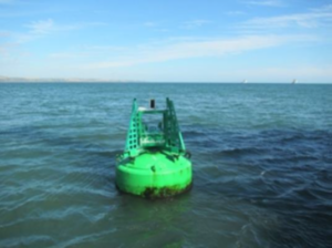 A green navigation buoy floats on calm blue-green water under a clear sky. The coastline is visible in the distance, with two small boats on the horizon.