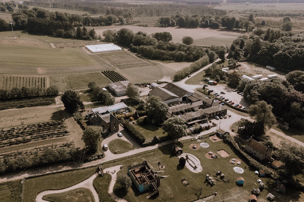 Aerial view of a rural area with buildings, car parks, playgrounds, gardens, farmland, and trees. Cars are parked along roads, and open fields stretch into the distance. The weather appears clear and calm.