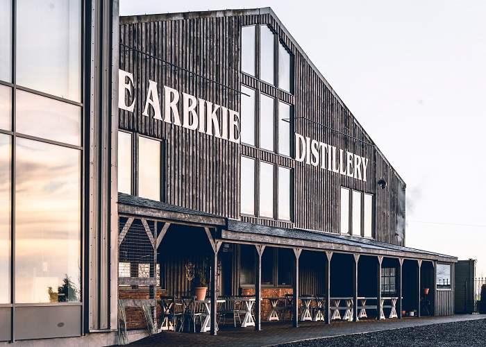 A wooden building with large windows and a wraparound veranda displays the words Arbikie Distillery in white letters. Outdoor seating and potted plants line the veranda. The sky is clear.