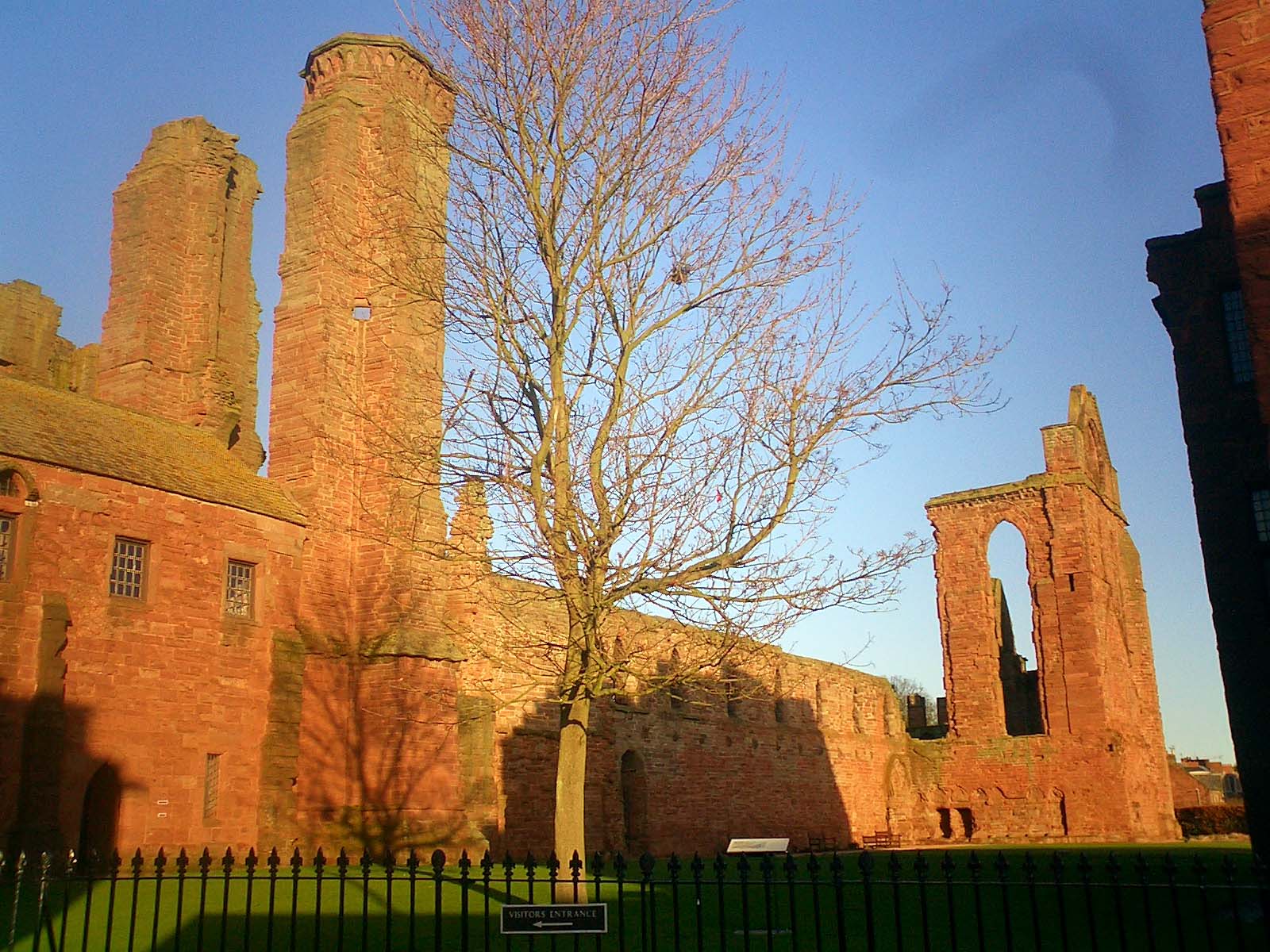 Red sandstone ruins of an old building with tall chimneys and archways, a leafless tree in front, black metal fence in the foreground, all under clear blue sky.