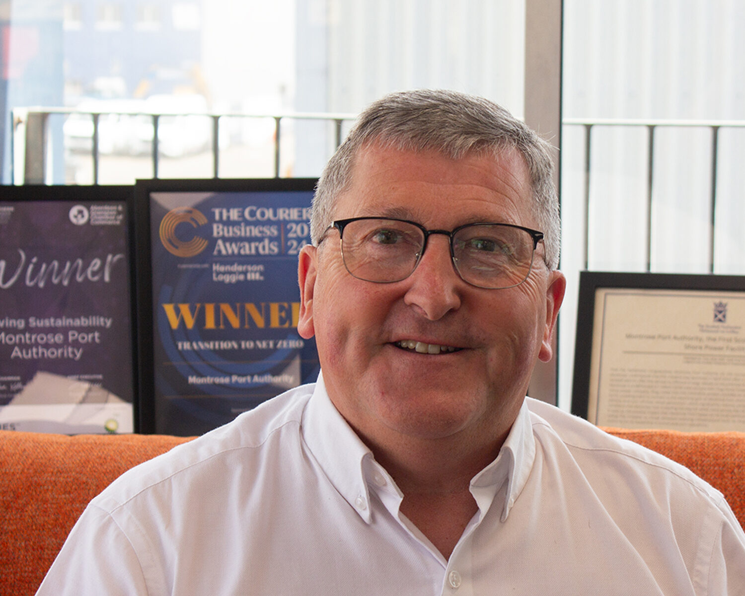 A man with short grey hair and glasses is sitting on an orange sofa, wearing a white button-up shirt. Award plaques and certificates are displayed on a shelf in the background.