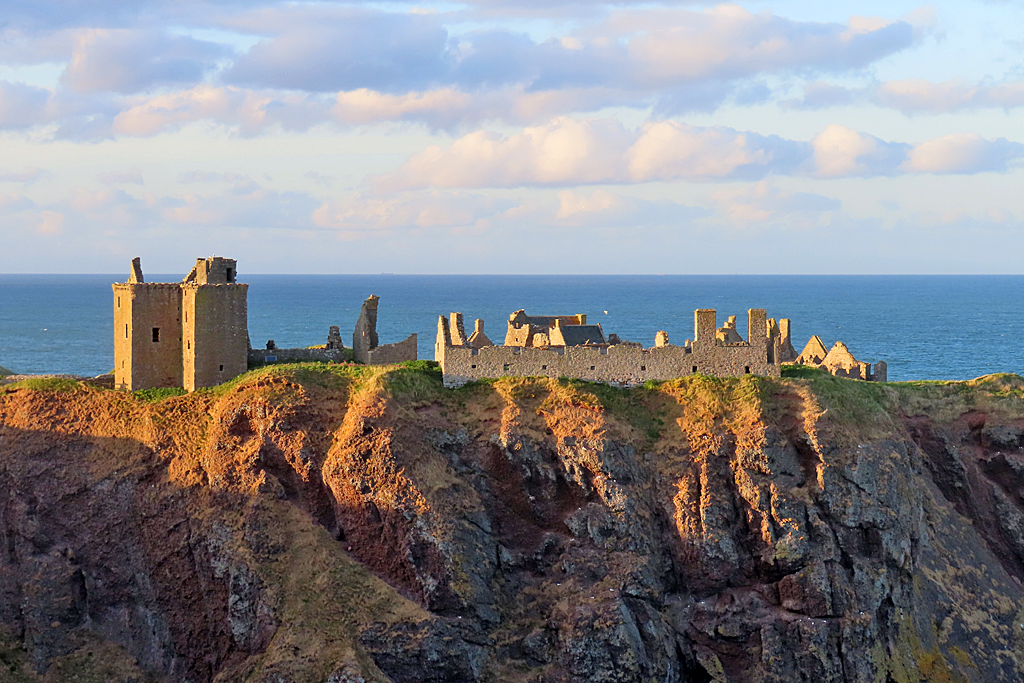Ruins of a stone castle sit on a grassy cliff overlooking the sea, with scattered clouds in the sky and sunlight casting shadows on the rocky cliff face.