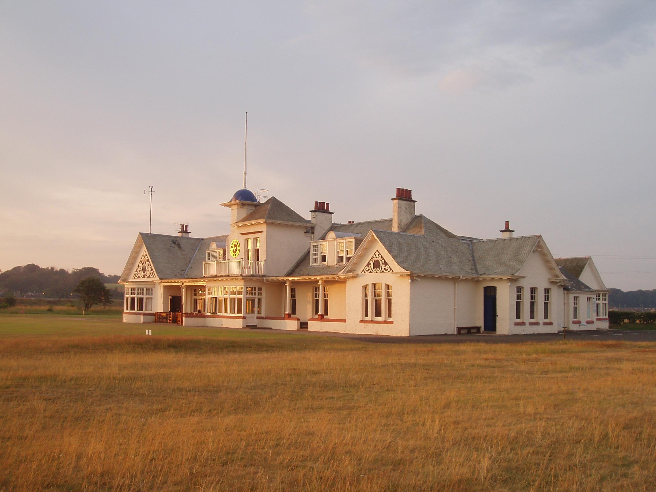 A large, white, historic-style building with a clock on the front stands on a grassy field under a sky with scattered clouds. The structure features multiple chimneys, gabled roofs, and decorative trim.