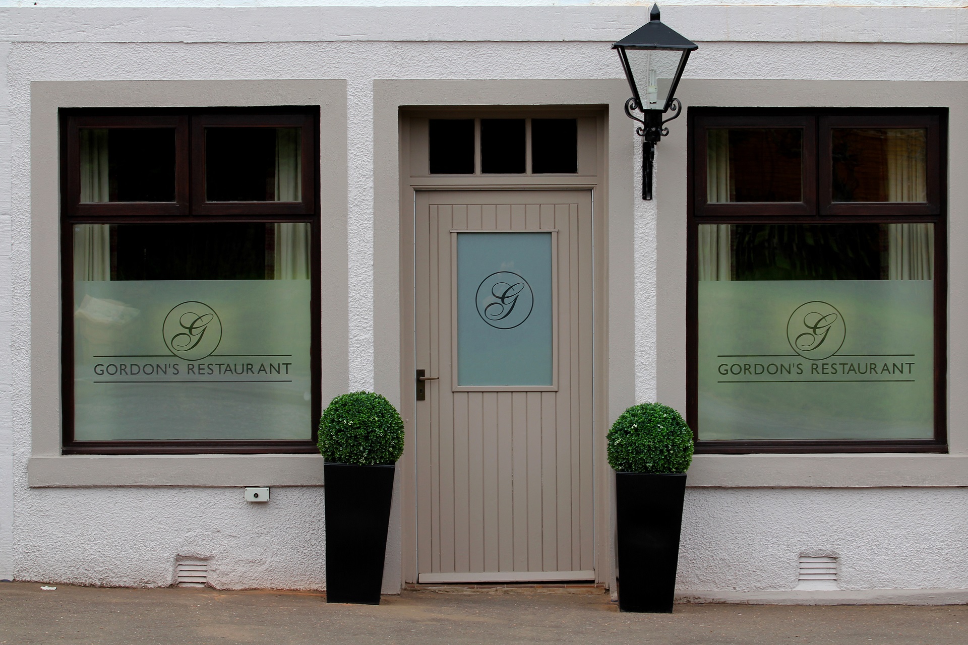The image shows the front of Gordon’s Restaurant, with a central beige door, two large windows labelled Gordons Restaurant, two round topiary plants in black pots, and a black wall lantern above the door.