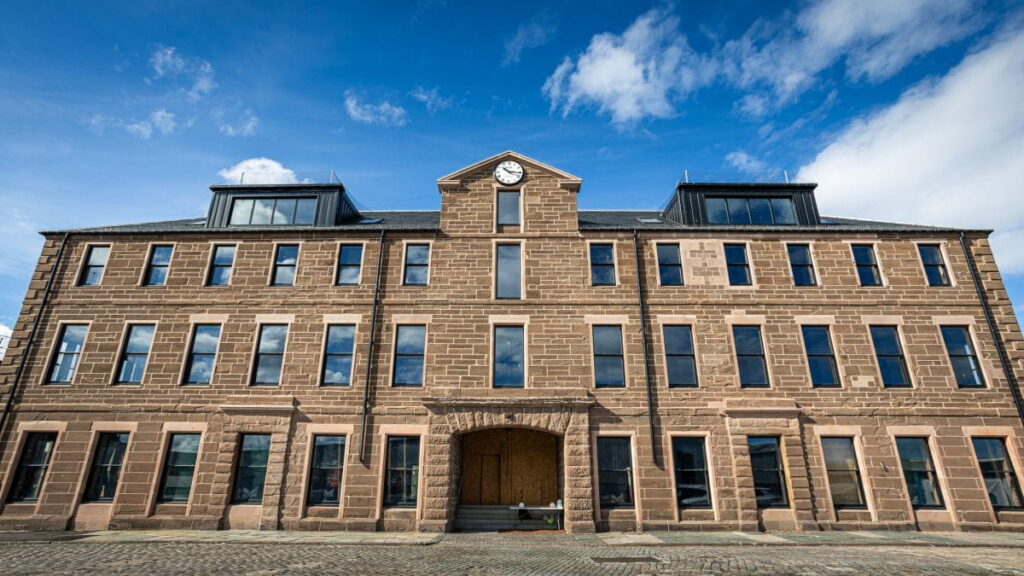 Three-storey stone building with many windows, a central arched entrance, and a small clock above the door—this is the Montrose Port Skills Academy, standing proudly under a blue sky with scattered clouds. Cobbled ground stretches in front of the structure.
