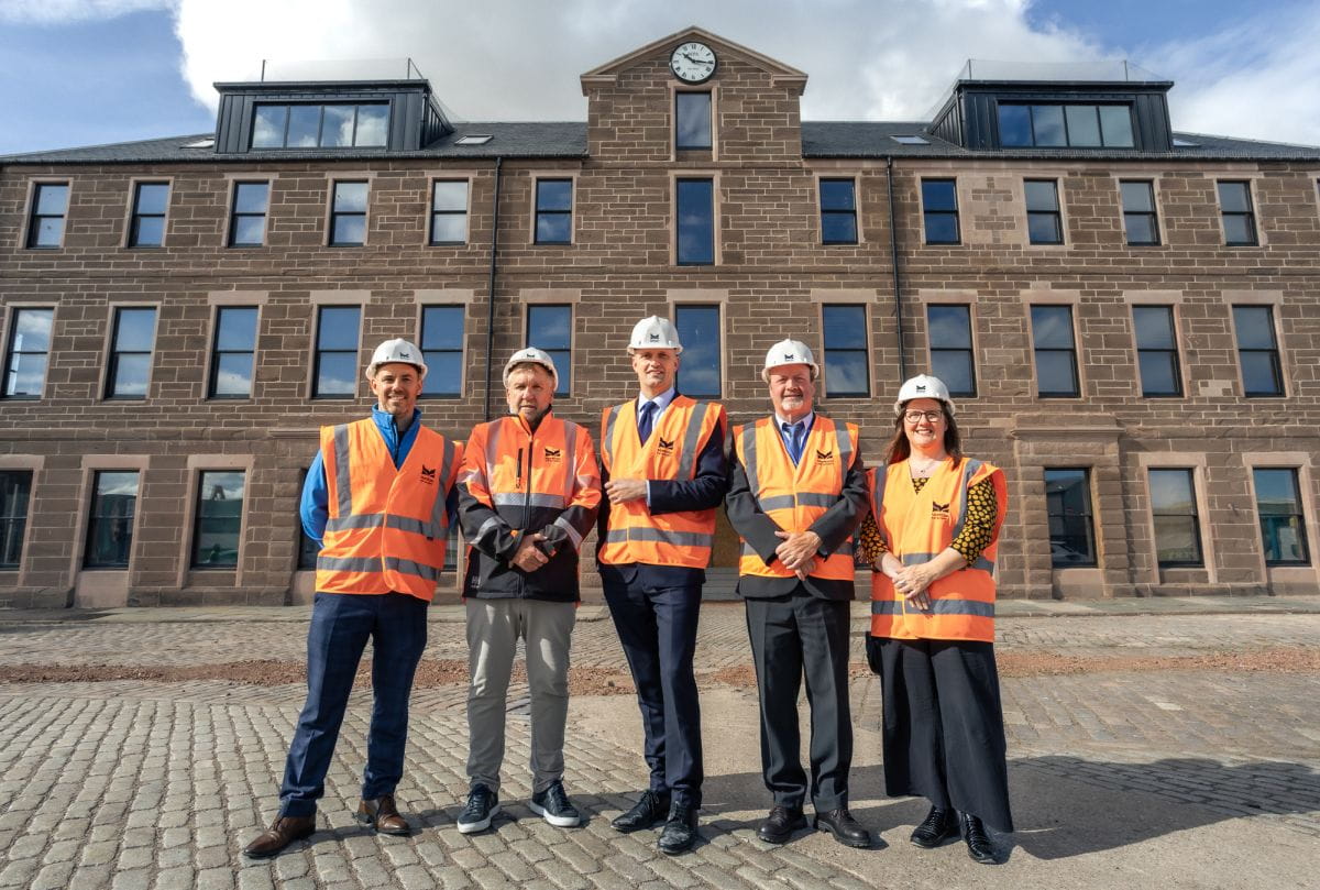 Five people wearing orange safety vests and white hard hats stand on a cobbled surface in front of a large stone building with a clock above the entrance. The group, participating in training with the Montrose Port Skills Academy, gathers under the partly cloudy sky.