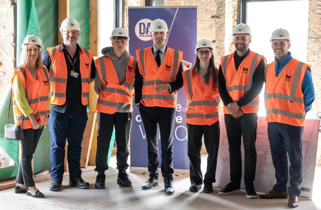 Seven people wearing hard hats and orange safety vests stand in a row inside a construction site. They are positioned in front of a banner for the Montrose Port Skills Academy and construction plans, with exposed walls and windows in the background.