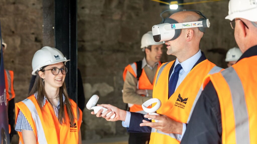 People wearing hard hats and orange safety vests are at a construction site. One person in a suit uses a virtual reality headset and controllers, while others observe and interact—a hands-on training session organised by the Montrose Port Skills Academy.