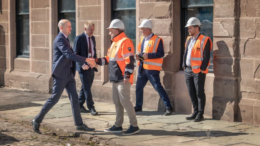 Two men, one in a suit and one in a construction jacket and helmet, shake hands outside a stone building—perhaps marking a partnership through the Montrose Port Skills Academy. Three other men in safety gear stand nearby, two wearing orange vests and one holding a camera.