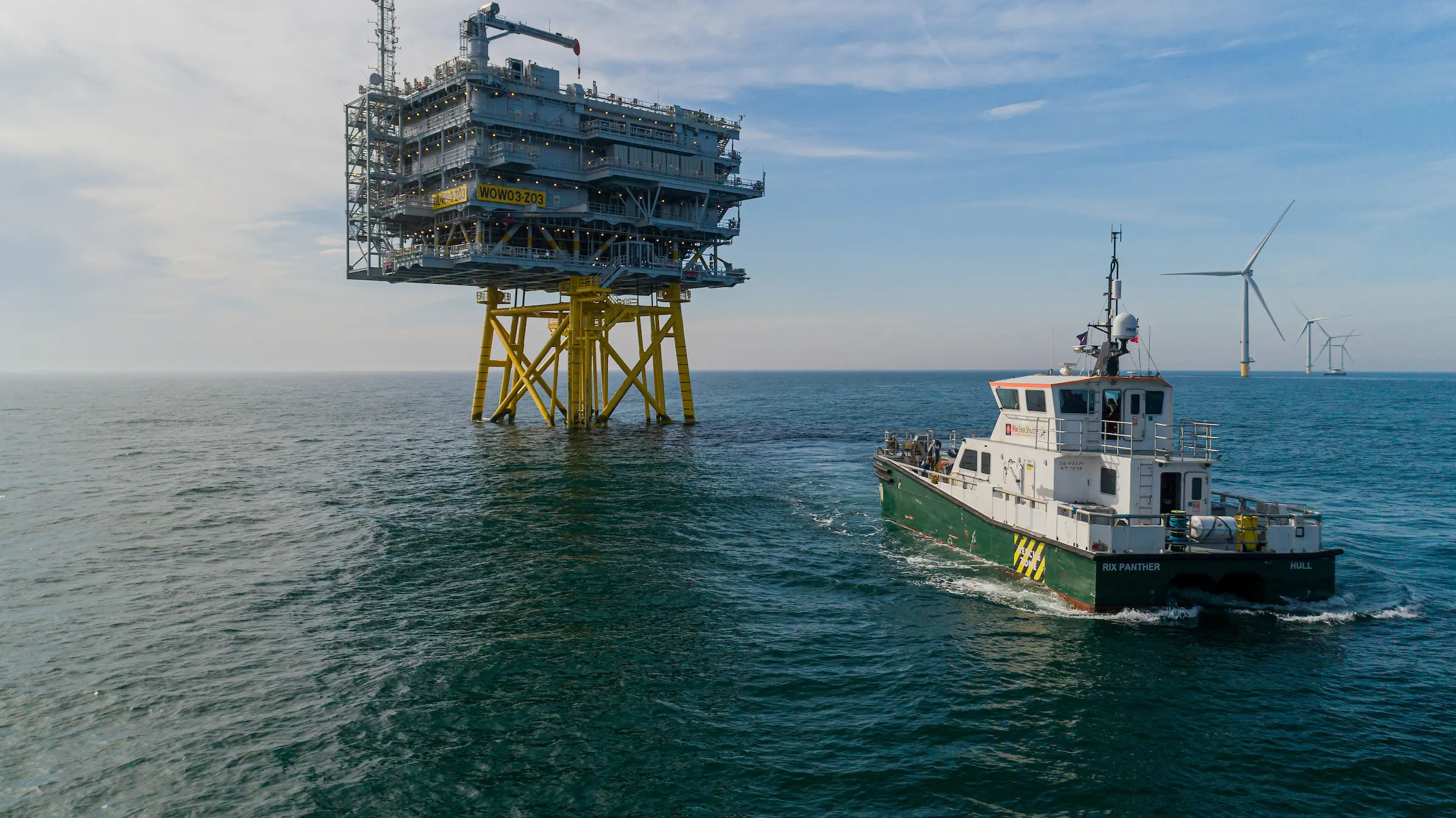 A large offshore oil platform stands on yellow supports in the sea. A green and white service boat approaches. Wind turbines are visible in the distant background under a partly cloudy sky.