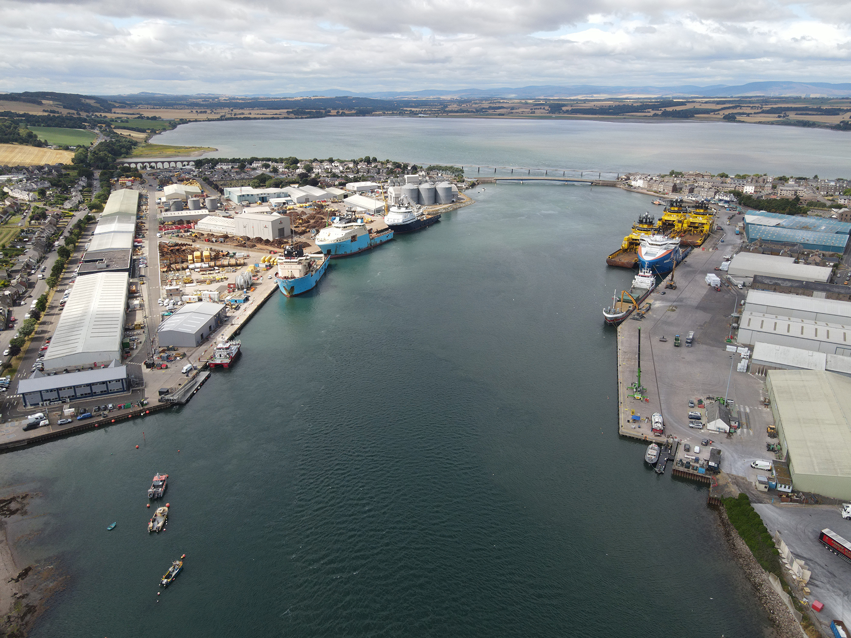Aerial view of a port with several ships docked along both sides, surrounded by industrial buildings and warehouses. Open water leads to a distant bridge, with farmland and hills visible in the background under a cloudy sky.