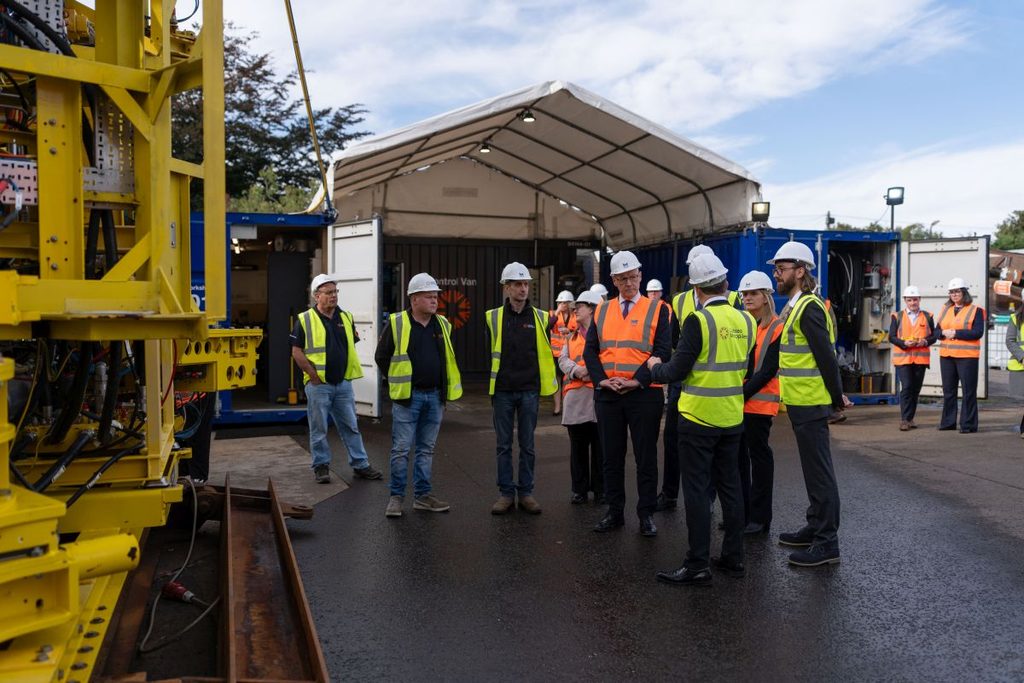 A group of people in safety vests and hard hats stand near a large yellow industrial machine at the Berwick Bank B site, having a discussion outdoors by temporary white marquees and equipment trailers on a tarmacked surface.