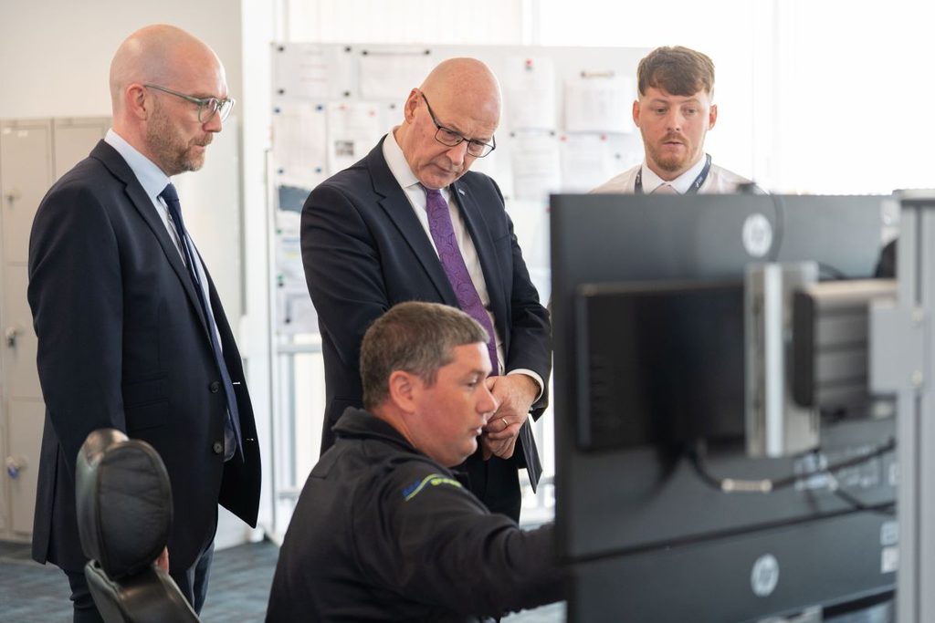 Four men in business attire stand around a seated colleague at a computer workstation, possibly discussing Berwick Bank B. Documents are posted on the wall in the background.