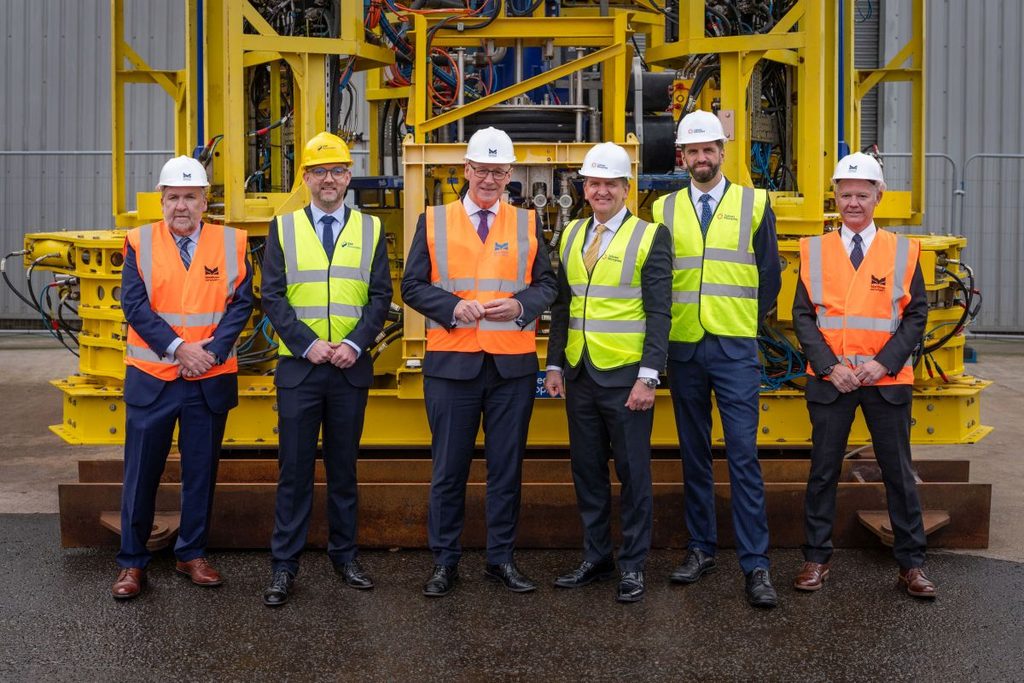 Six men stand in front of industrial machinery, wearing hard hats and safety vests. Three wear orange vests, two wear yellow vests, and one is without a vest. They stand on wet pavement, posing for a group photo.