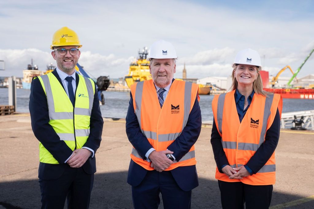 Three people stand outdoors at Berwick Bank B port. Two wear orange safety vests and white hard hats, while one wears a yellow vest and yellow hard hat. Ships and industrial equipment are visible under a partly cloudy sky.