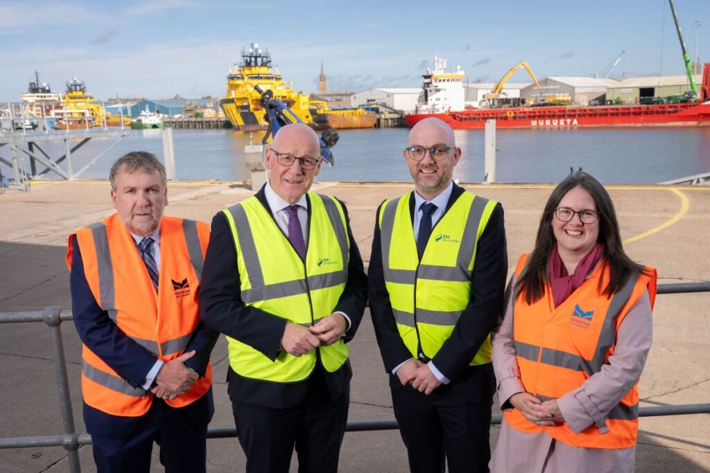Four people in safety vests and business attire stand on a quay, posing for a photo. Large ships, cranes, and port equipment are visible in the background under a partly cloudy sky.